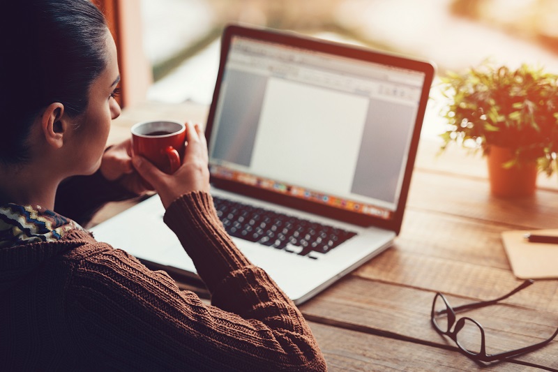 Close-up image of pensive young woman holding coffee cup and looking away while sitting at her working place at the rough wooden table.